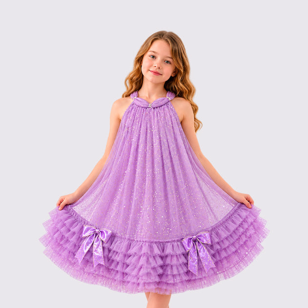 A young girl with wavy light brown hair wearing a lavender purple glitter halter dress with a tiered tulle skirt and matching bow details, standing against a solid light gray background.